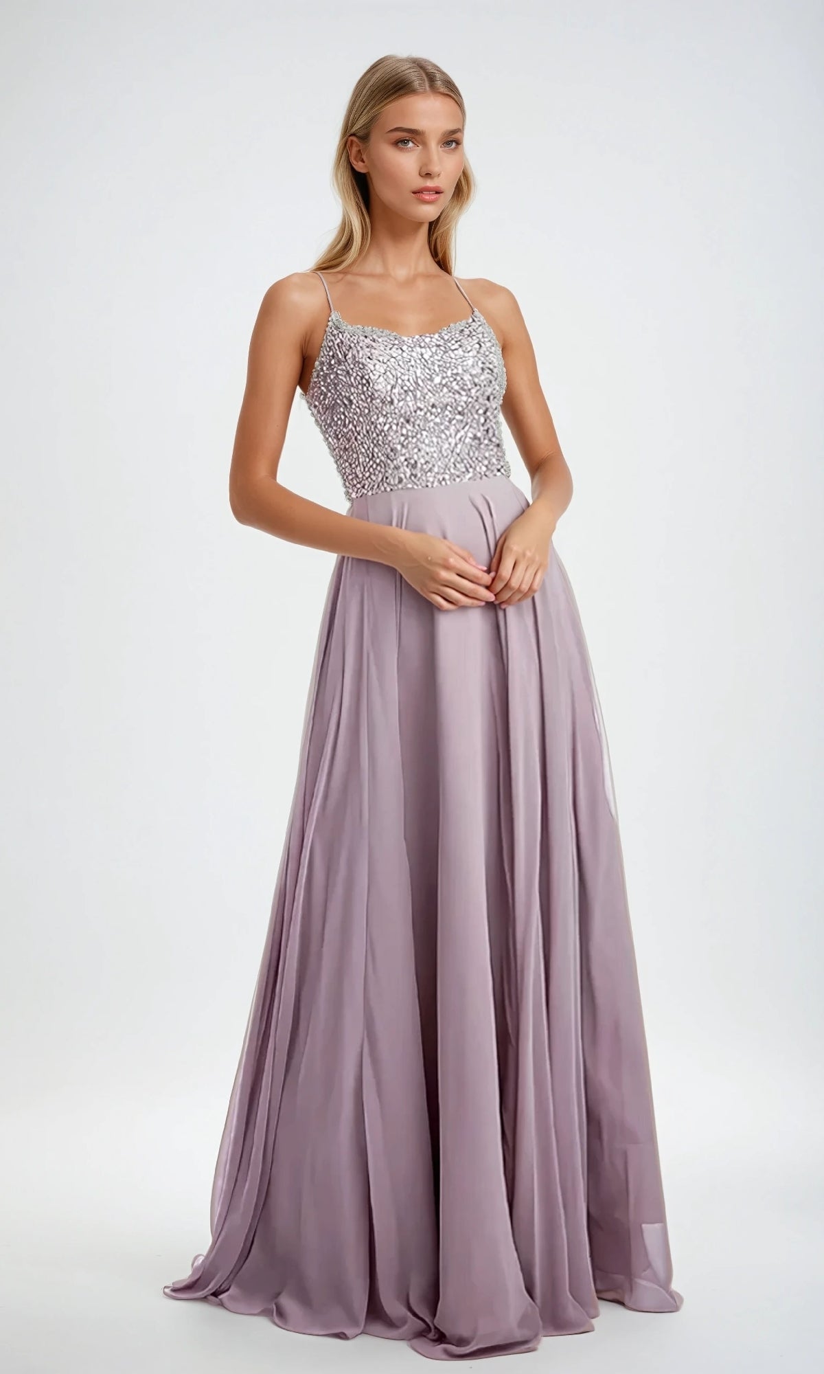 A woman stands wearing the Juliet 210 Long Prom Dress, featuring a floor-length lilac design with a sequined bodice, thin straps, and a flowing A-line skirt against a plain background.